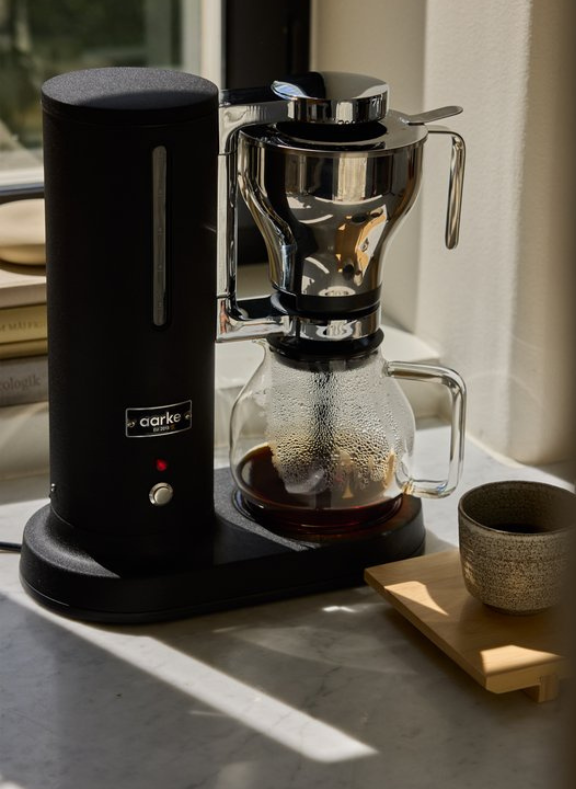Aarke Matte Black coffee maker with glass carafe on a marble surface, sunlight casting shadows.