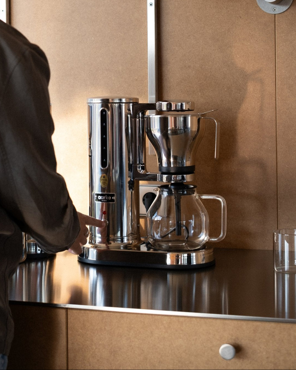 Person using an Aarke stainless steel coffee maker on a modern kitchen counter.