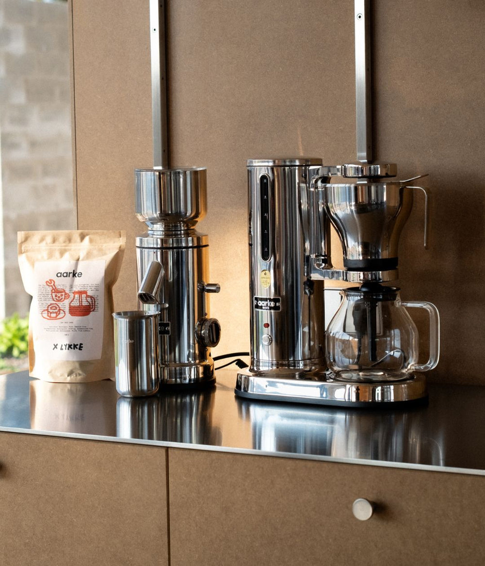 Aarke Coffee Grinder, Coffee Maker, and accessories on a modern kitchen counter alongside Aarke x Lykke coffee beans.