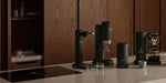 Collection of aarke products in matte black on a kitchen counter with wooden cabinets in the background.