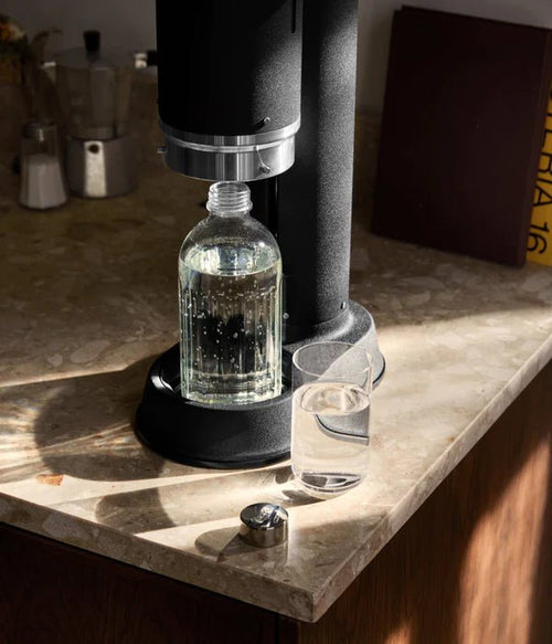 The Aarke Carbonator Pro in matte black placed on a sunlit kitchen counter, next to a filled glass and kitchen accessories.
