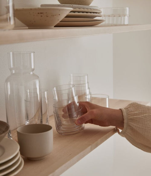 A hand reaching for stacked Aarke crystal glasses on a wooden shelf in a softly lit kitchen setting.