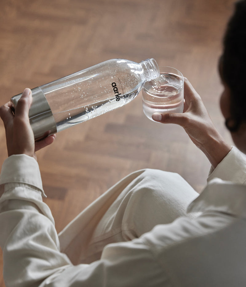 A person pouring water from an Aarke PET Water Bottle for Carbonator 3 into a glass, with a wooden floor visible in the background.