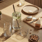 A table with food has a full nesting carafe in the center. The mixing spoon is inside along with water, cucumber, and ice. Beside the carafe are a set of nested glasses.