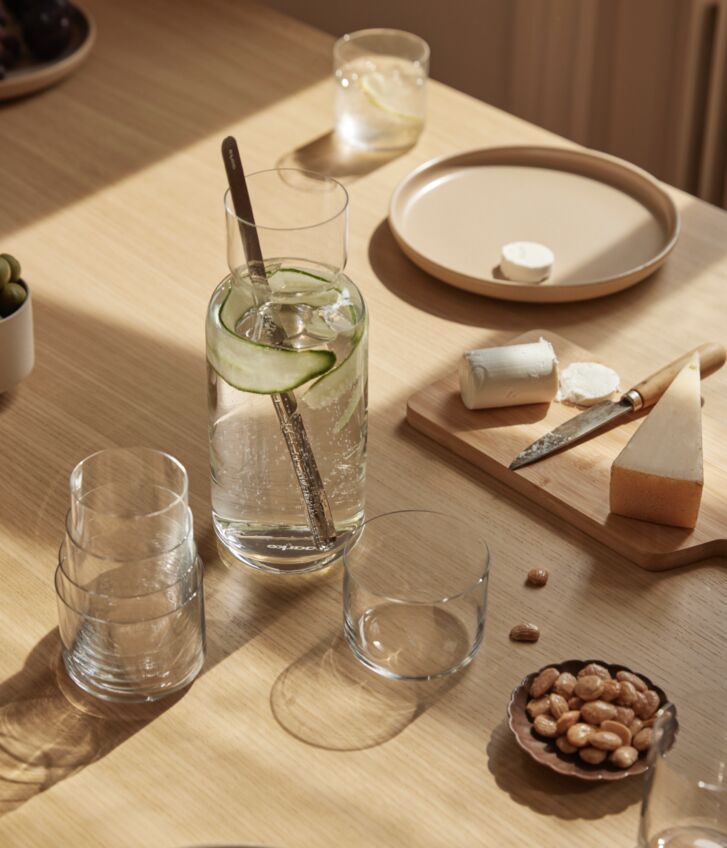 A table with food has a full nesting carafe in the center. The mixing spoon is inside along with water, cucumber, and ice. Beside the carafe are a set of nested glasses.